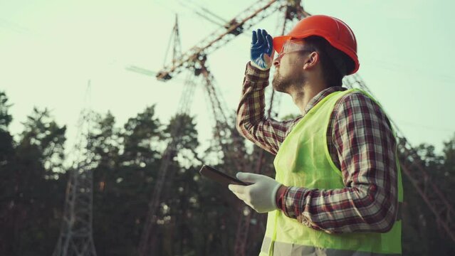 Energy industry engineer works at power plant with tablet in protective vest and hard hat. Engineer inspecting high voltage power grid by using tablet. Electric poles and power lines in background