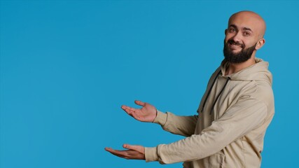 Smiling person advertising something on camera, pointing to left or right sides in studio. Middle eastern guy showing advertisement for marketing campaign, blue background pose. Camera 1.