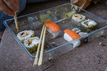Young woman wearing jeans is sitting on the stone steps eating sushi rolls out of opened half-empty...