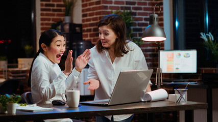 Jubilant businesswoman and coworker celebrating finalizing deal with customers after working together on laptop. Excited sales consultants doing high five after getting quality leads