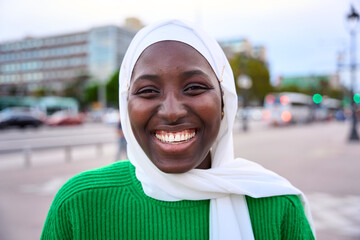 Portrait of black muslim woman very happy, smiling and looking at camera. An African student sincerely smiles while standing outdoors. The girl is wearing a white hijab and green t-shirt