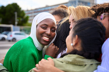 Portrait of black Muslim woman looking smiling at camera surrounded in an embrace by multiracial female people. African girl student in white hijab happy with her friends standing together outdoor