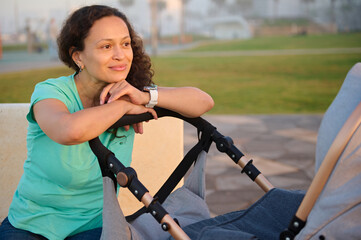 Fototapeta premium Latina American happy beautiful woman, young mum smiles looking aside, putting her hands on the baby pram with her sleeping child, enjoying walking at sunset. People. Maternity leave lifestyle concept