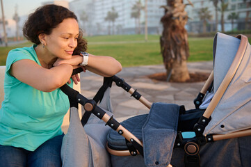 Obraz premium Beautiful multi ethnic young woman, mother 40s, smiling while admiring her baby sleeping in the baby pram, enjoying a happy weekend outdoors at sunset. People. Lifestyle. Leisure activity
