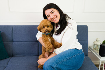 Cheerful young woman cuddles with her adorable brown poodle on the sofa in a bright, cozy living...