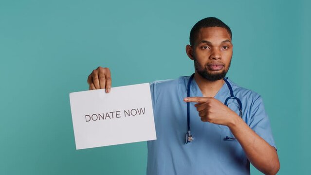 Portrait of smiling medic holding donate now sign, inviting people to provide aid for those in need, studio background. Healthcare employee requesting philanthropic donations for patients, camera A
