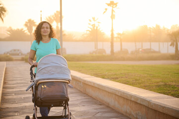 Happy smiling young woman, mother looks at camera while pushes silver gray baby carriage. Infant...