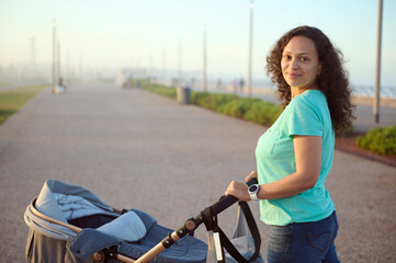 Young happy Latina woman 40s, loving mother looking at camera, pushing a baby pram while walking on...