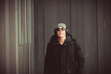 Portrait of Handsome Smiling Young Man with Woolen Hat, Sunglasses and Earphones on the Street Listening to Music