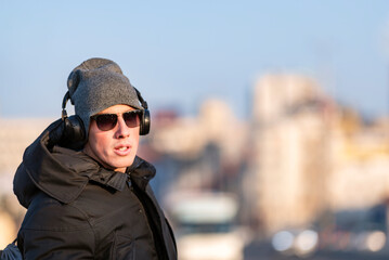 Portrait of Handsome Smiling Young Man with Woolen Hat, Sunglasses and Earphones on the Street Listening to Music