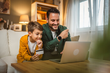 Son sit on sofa with father and hold credit card buy online on laptop