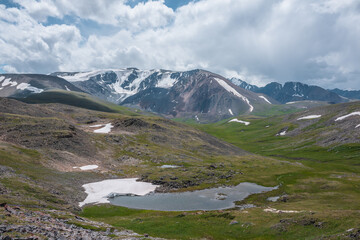 Beautiful small lake with snowfield among green grassy hills and rocks with view to snow-capped...