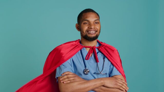 Smiling male nurse portraying superhero wearing red cape, isolated over studio background. Portrait of cheerful heroic medic posing as hero in costume with mantle, camera A
