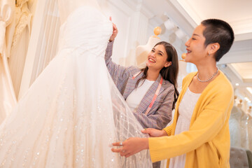 Naklejka premium Asian bride is smiling while choosing wedding dress in modern wedding salon, Attractive designer girl using tape meter fitting on wedding dress at wedding studio.
