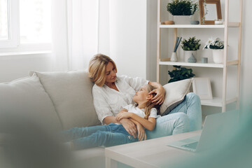 Mother and child enjoying quality time on the couch, laptop open on the table, bookshelf filled with books in the background