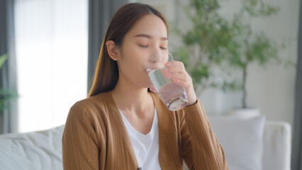 Happy young Asian woman drinking water while sitting on the sofa in the living room