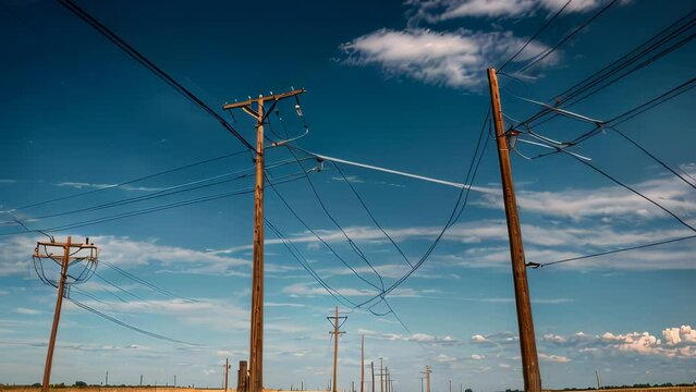 Video animation of electricity poles stretching across the landscape. Against a clear blue sky with sparse clouds, these wooden and metal structures stand tall, their wires crisscrossing the frame