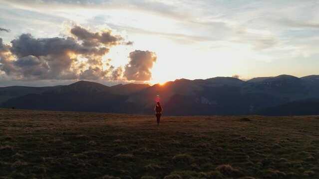 Aerial drone back follow shot of young woman hiker walking on a hill with mountains and sunset light in the background