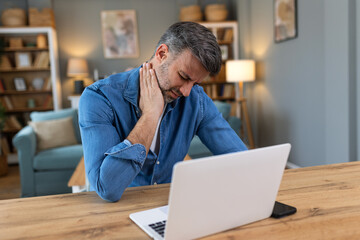 Businessman feeling pain in neck after sitting at the table with laptop. Tired man suffering of office syndrome because of long hours computer work. He is massaging his tense neck muscles