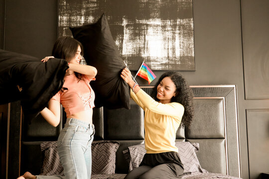 Happy mixed-race lesbian couple having fun during a pillow fight on a bed mattress. Two cheerful teenage lesbians laughing and playing funny active games in the bedroom, LGBTQIA pride month concept.