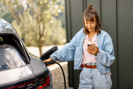 Young woman plugs a charger into electric vehicle standing with smart phone near her house outdoors. Concept of modern technologies, EV cars and sustainable lifestyle