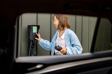 Woman switches on car charging station while charging her electric vehicle near home. Concept of smart technologies for comfort living, EV cars