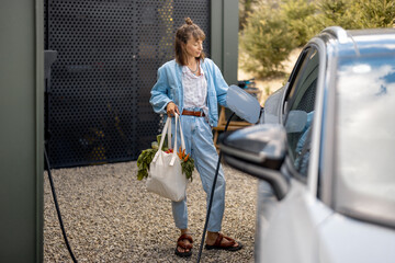 Young woman plugging a charger into electric vehicle, carrying bag with fresh vegetables near her...