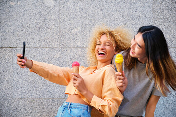Two multiracial happy friends with ice creams taking a selfie in summer at street.