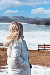 Cute blonde girl in warm clothes while walking, standing against the backdrop of a frozen lake. 