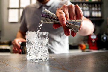 A person pours a clear liquid from a jigger into a glass filled with ice cubes, likely preparing a beverage