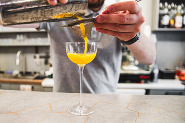 Bartender pouring a vibrant orange cocktail into an elegant glass, with a twist of lemon zest being...