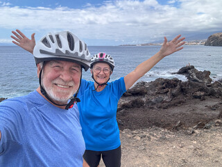 Cheerful active couple of seniors biking with electric bicycles at sea taking a memory photo enjoying healthy lifestyle in retirement. Horizon over water