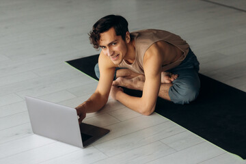 Handsome young man looking at laptop screen and stretching. Man having fitness class online in the...