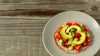 Red corn tortilla topped with Pico de Gallo salsa and avocado slices on a plate. Rustic wooden table with space for text. Healthy vegan Mexican food.