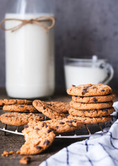 Galletas con chispas de chocolate apiladas sobre una rejilla de enfriamiento en una cocina rústica. En el fondo una botella y una taza de leche. Iluminación suave en entorno acogedor y hogareño.