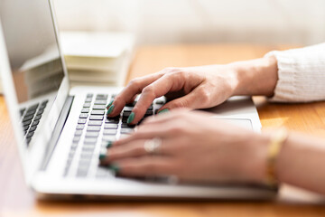 Primer plano de manos femeninas con uñas pintadas de verde, escribiendo en un teclado de portátil sobre un escritorio de madera, con una agenda abierta al fondo en un entorno de oficina.






