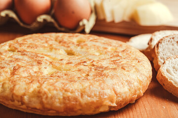 Tortilla Española de patatas entera junto a unas rodajas de pan y de fondo los ingredientes, huevos y patatas.