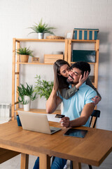 Young happy couple online shopping while using laptop and credit card at home. Caring wife kissing...