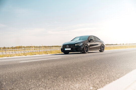 Photo of a black Mercedes-Benz AMG CLA 45 S 4MATIC+. They show details of the car from the outside. Rims, emblems, headlights, lamps in a sporty style. The model stands in the middle of highway.