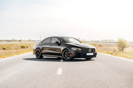 Photo of a black Mercedes-Benz AMG CLA 45 S 4MATIC+. They show details of the car from the outside. Rims, emblems, headlights, lamps in a sporty style. The model stands in the middle of highway.