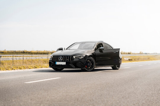 Photo of a black Mercedes-Benz AMG CLA 45 S 4MATIC+. They show details of the car from the outside. Rims, emblems, headlights, lamps in a sporty style. The model stands in the middle of highway.