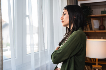 Young sad and depressed woman feeling regret and anxiety standing near window looking through....