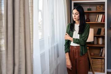 Young sad and depressed woman feeling regret and anxiety standing near window looking through....