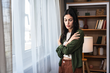 Young sad and depressed woman feeling regret and anxiety standing near window looking through....