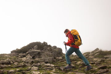 Naklejka premium A woman is hiking up a mountain with a yellow backpack. She is wearing a red jacket and blue pants. The sky is cloudy and the weather is cool