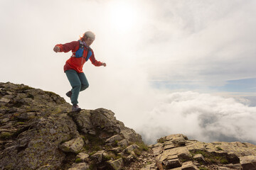 A woman is standing on a mountain top, wearing a red jacket and blue pants