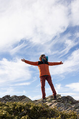 A man in an orange jacket stands on a rocky hillside, looking up at the sky