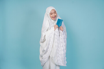 Fototapeta premium Happy young Asian Muslim woman wearing prayer gown holding passport isolated by blue background. Hajj concept.