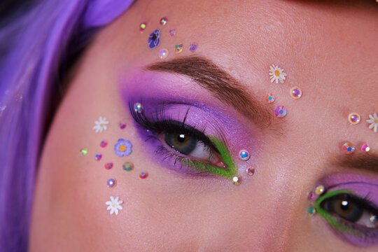 Stunning spring purple and neone green eye makeup with doll eyeslahes, flowers and perals. Girly makeup for spring and summer festivals. Close up on face.