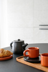 Ceramic teapot with tea, cups on wooden tray and fresh croissant on table
